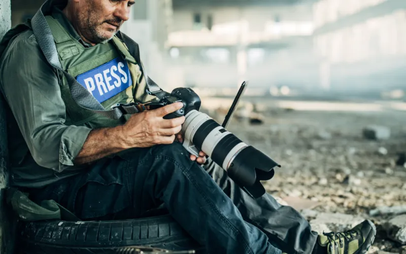 A male photojournalist in a "Press" vest examines his camera while seated on a tire in a dusty, abandoned building, conveying focus and determination.