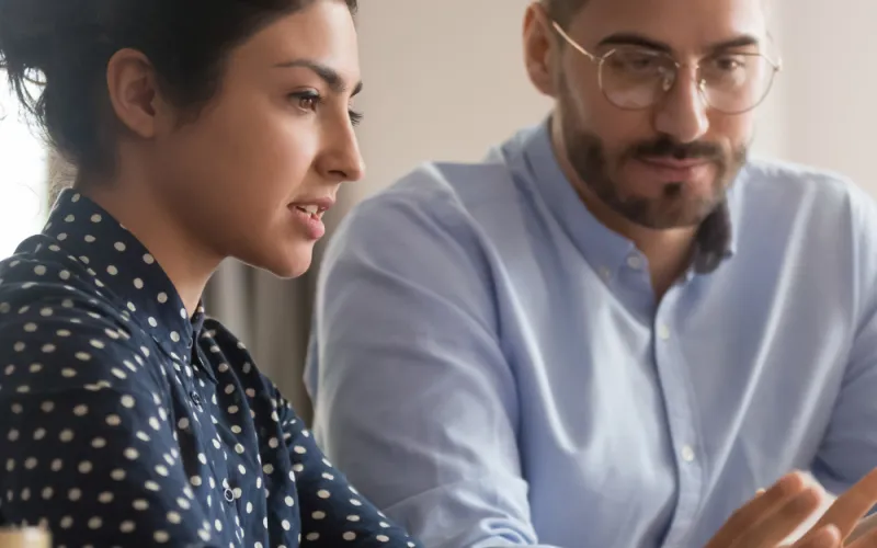 A woman in a polka dot blouse and a man in glasses and a light blue shirt are engaged in a focused discussion at a desk, conveying teamwork.