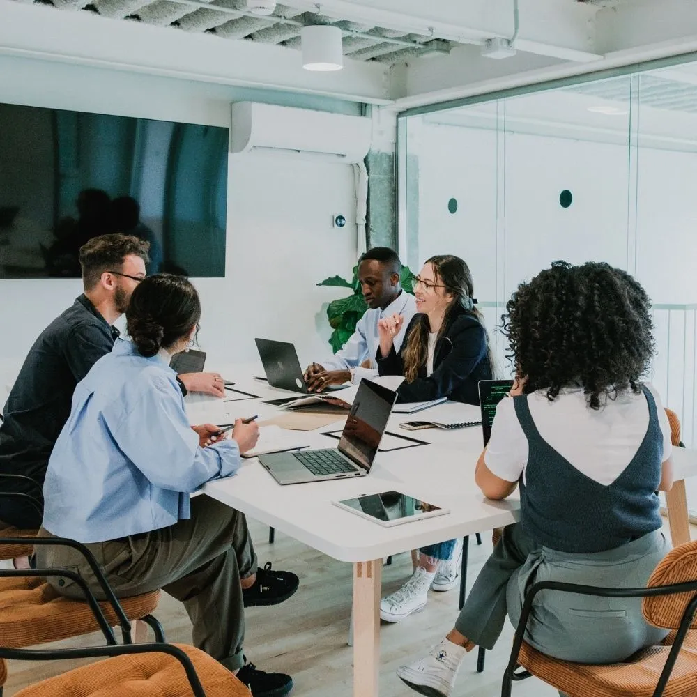 A diverse group of five people engaged in a team meeting around a table with laptops in a modern, bright office. They appear focused and collaborative.