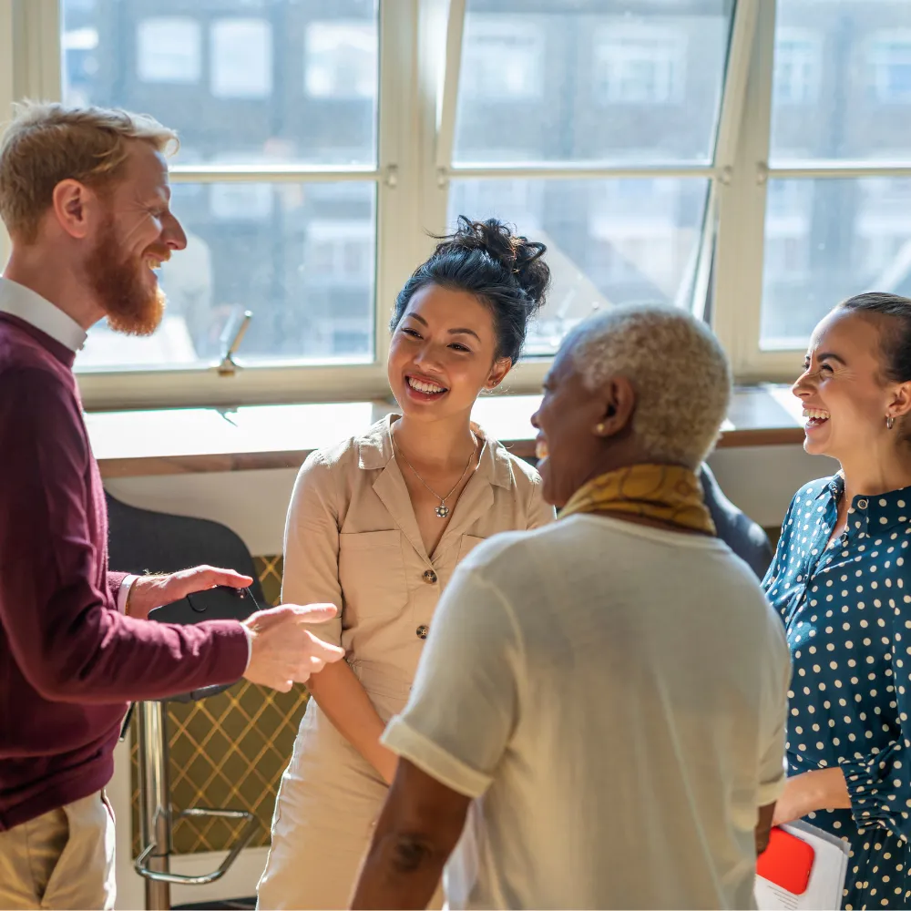 A diverse group of four people standing in a sunlit room, smiling and engaging in friendly conversation, conveying a positive and warm atmosphere.