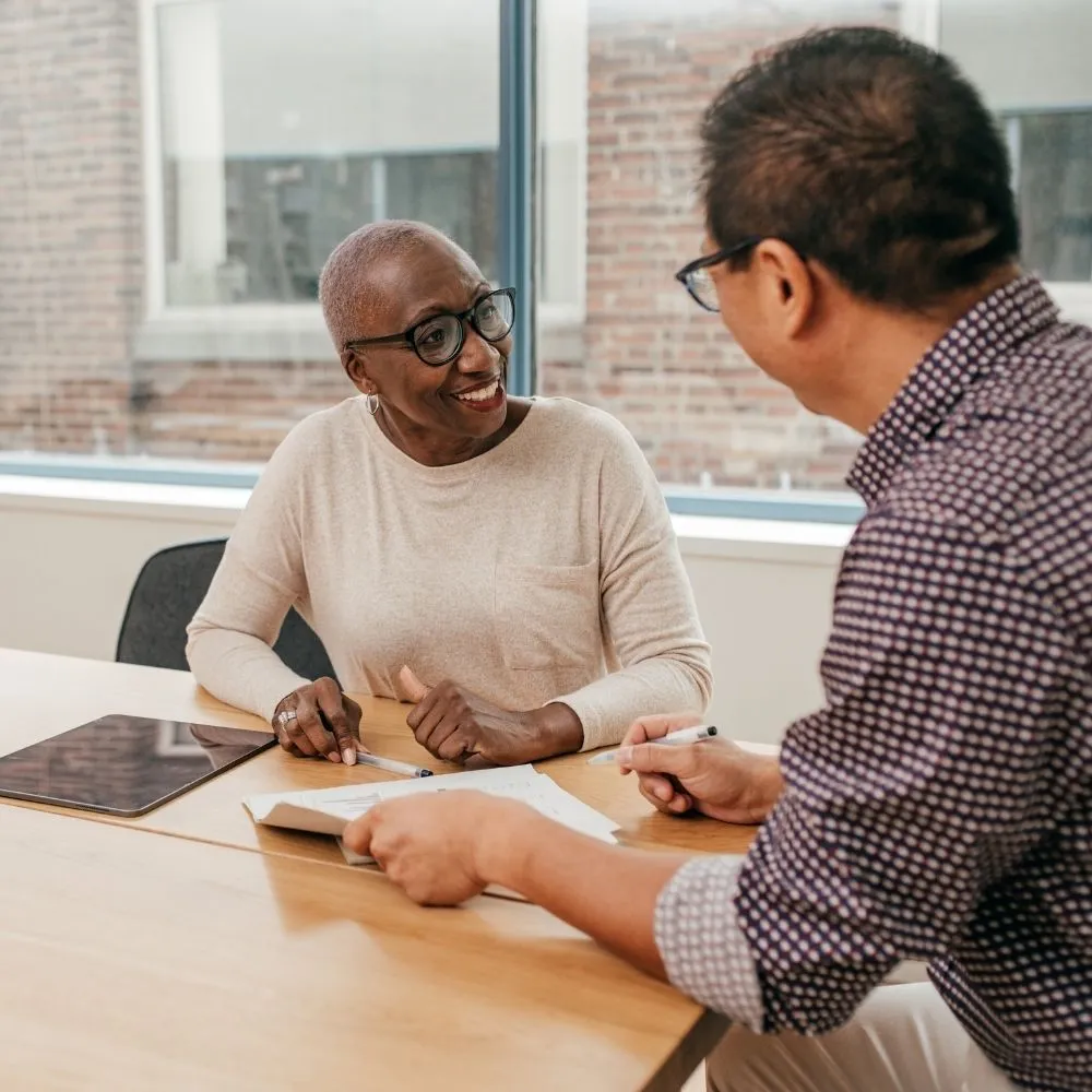Two people are seated at a table, engaged in a friendly discussion. Both are smiling, with documents and a tablet in front of them. Bright, casual office setting.
