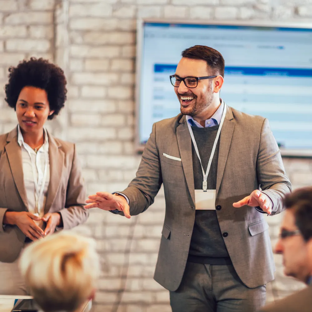 A man in a suit, smiling and gesturing energetically, leads a meeting. A woman beside him looks attentive. 