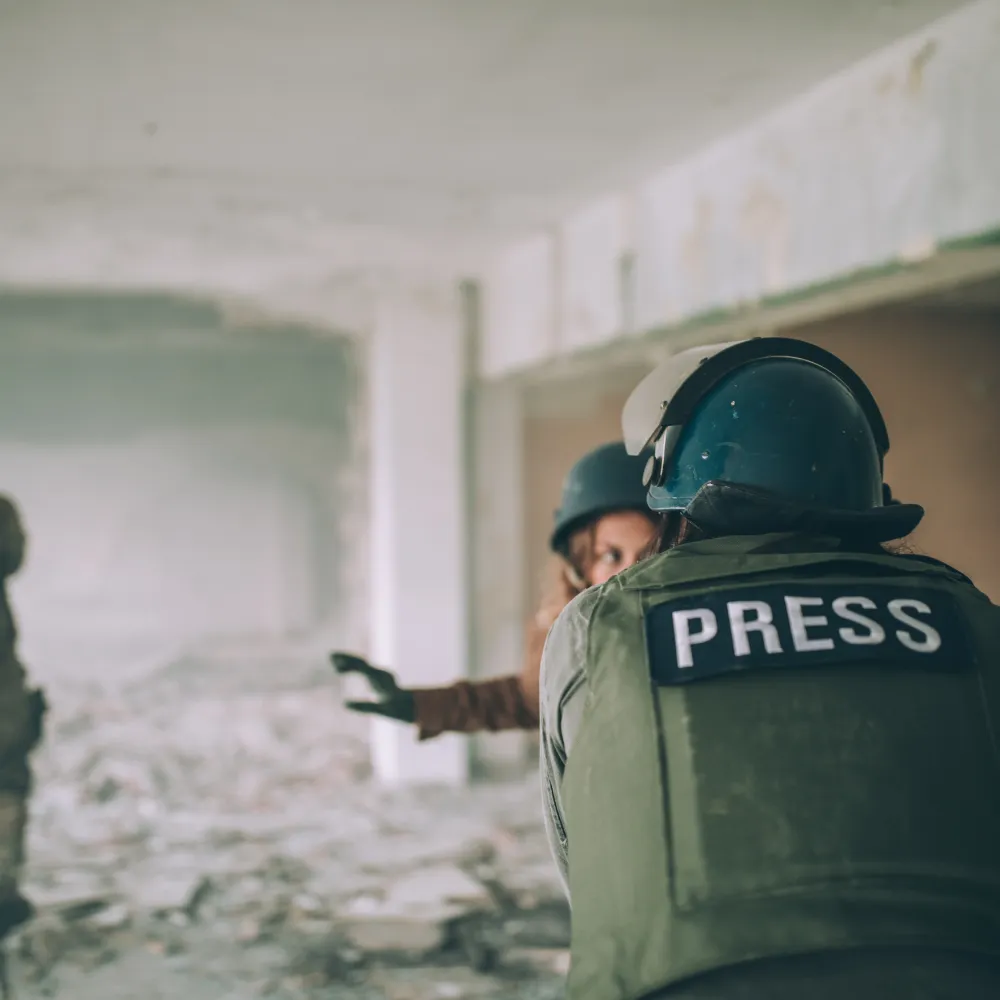 A press reporter in a helmet and vest labeled "PRESS" gestures urgently in a crumbling, debris-filled building, conveying a tense atmosphere.