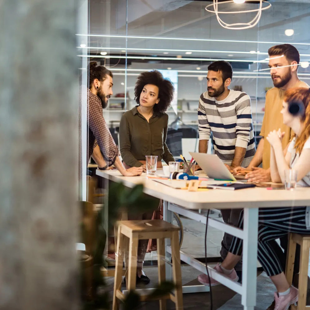 A diverse group of five young professionals in a modern office, engaged in a focused discussion around a table with a laptop and documents.