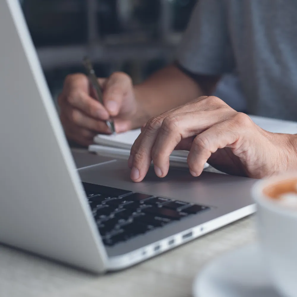 A person works at a laptop, typing with one hand and taking notes with a pen in the other, with a cup of coffee nearby, conveying focus and productivity.