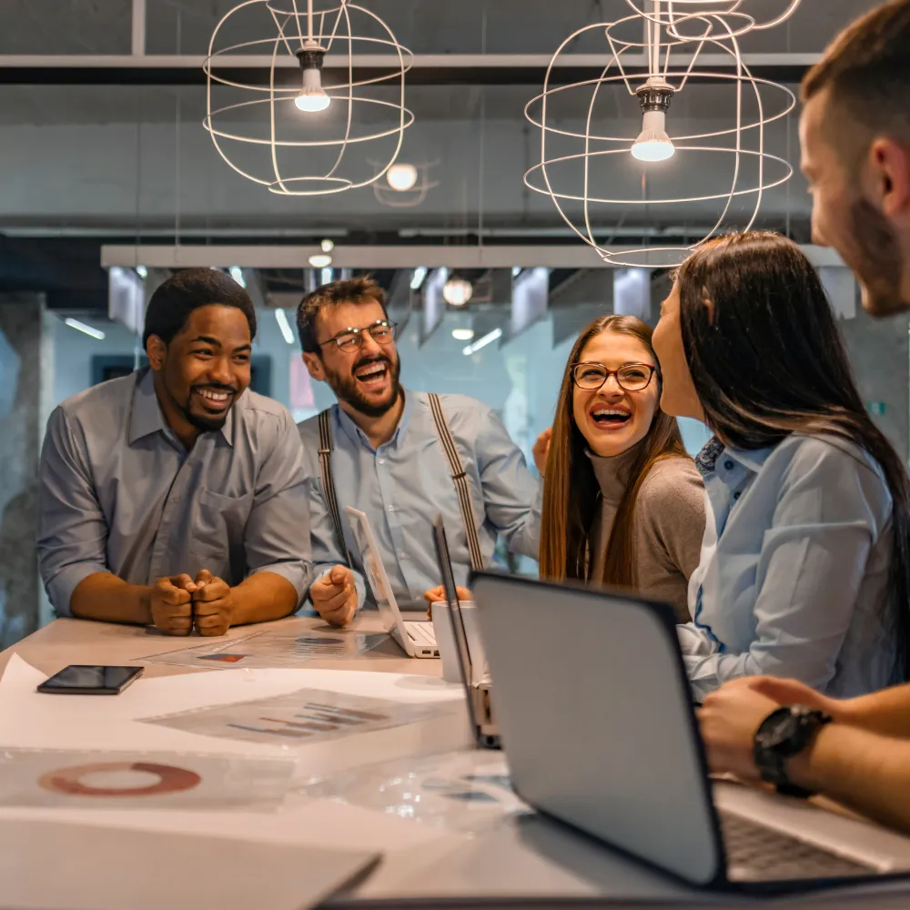 A diverse group of colleagues in casual attire are joyfully laughing around a table with laptops and papers in a modern, well-lit office setting.