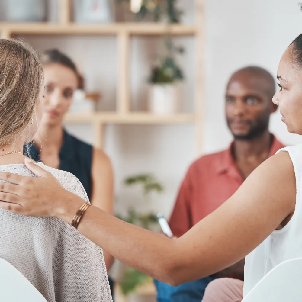 A supportive group setting with four people. One woman comforts another by placing a hand on her shoulder. The mood is empathetic and caring.