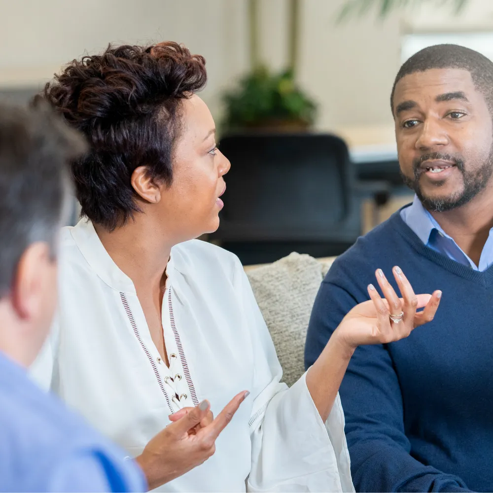 A woman in a white blouse and a man in a blue sweater engage in an active conversation, showing expressive gestures, conveying a collaborative tone.