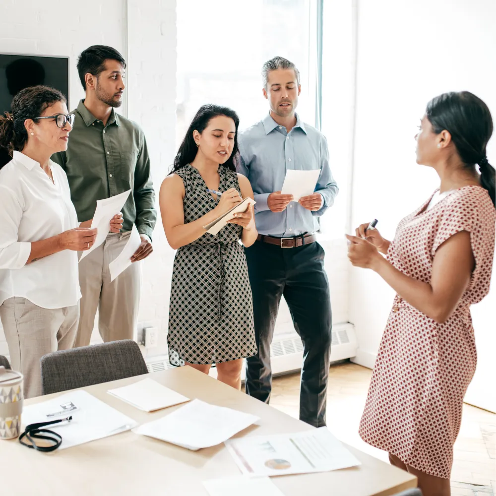 A group of five people stand in a bright office, engaged in discussion. They hold papers and a notebook, suggesting a collaborative meeting.