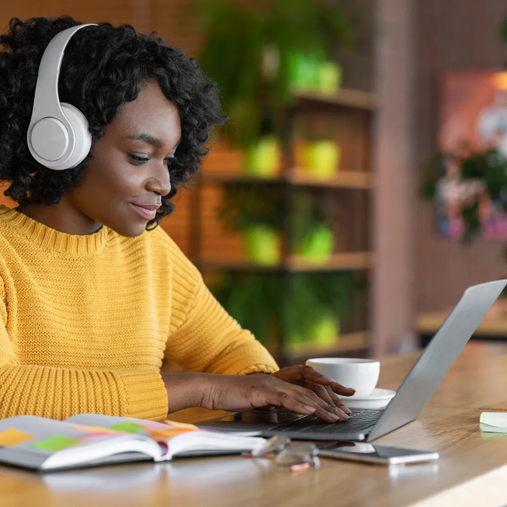 A woman in a yellow sweater sits at a table with a laptop, wearing white headphones. 