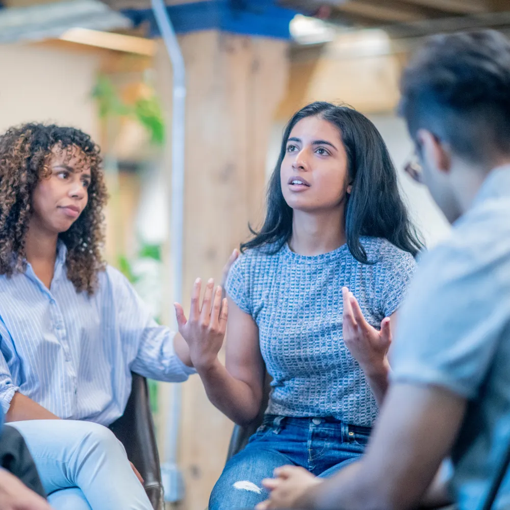 A woman gestures passionately while speaking in a group setting. Another woman listens intently, with a hand on her shoulder offering support. 