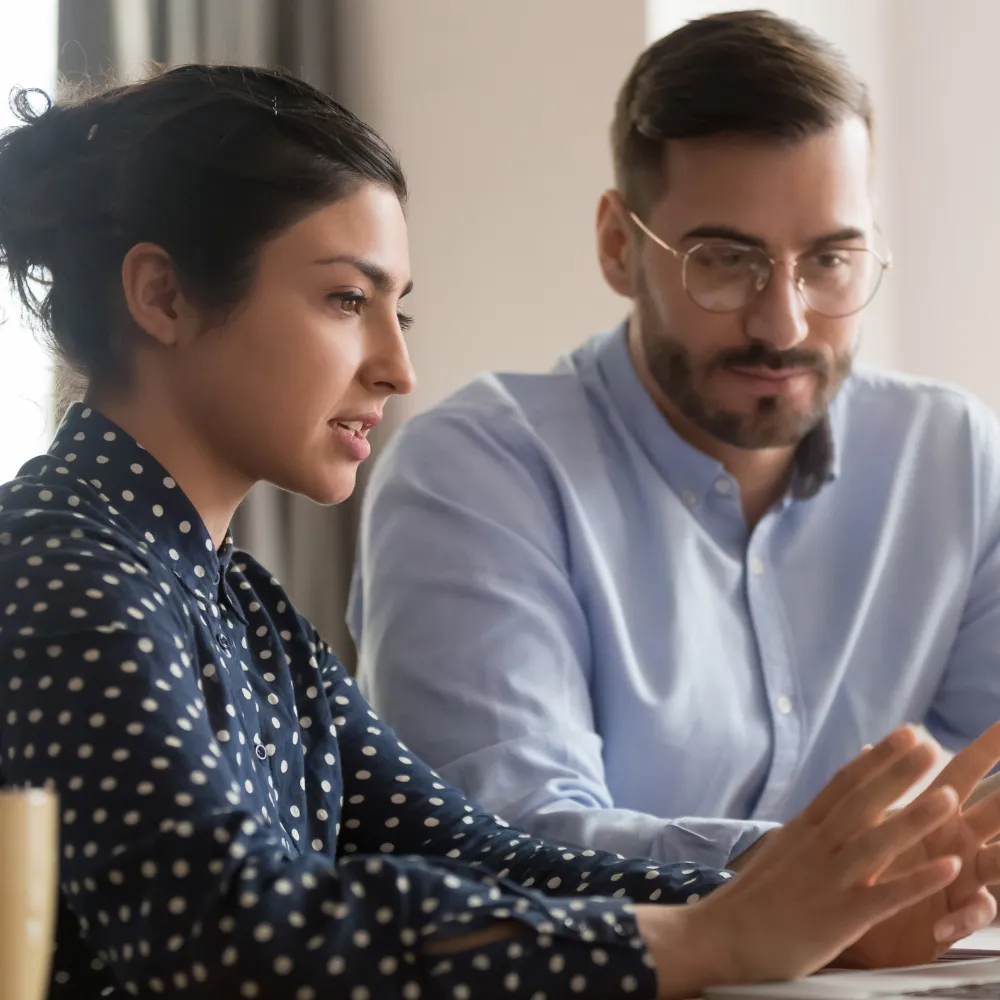 A woman in a polka dot blouse and a man in glasses and a light blue shirt are engaged in a focused discussion at a desk, conveying teamwork.