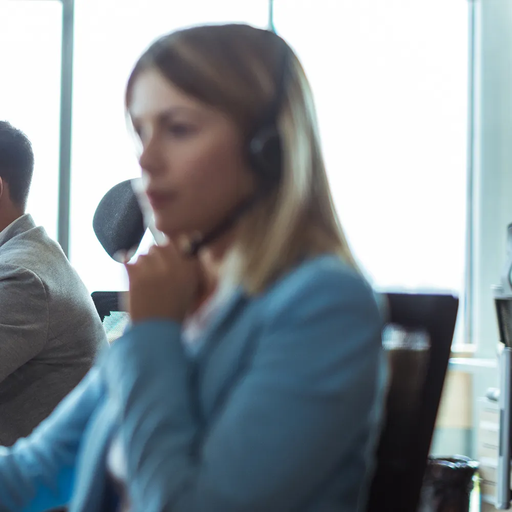 A woman in a blue blazer and headset sits in an office, focused on a call.