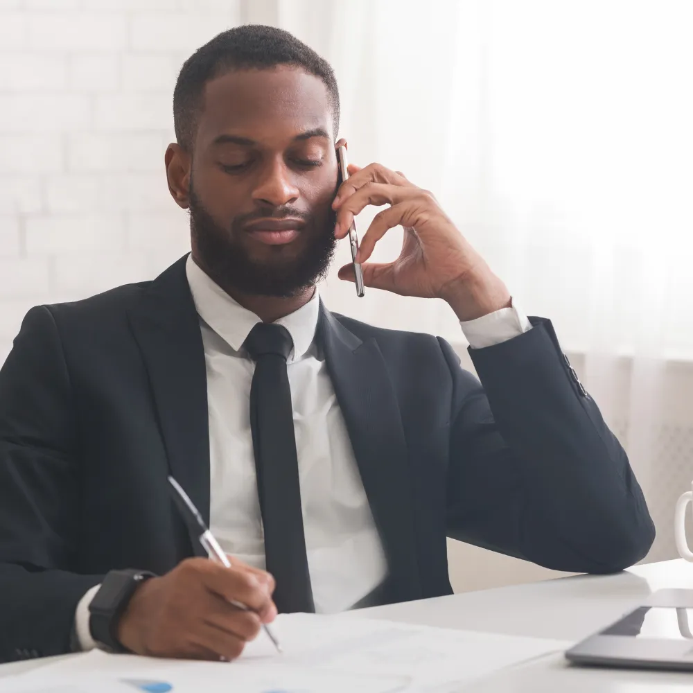 A man in a suit and tie sits at a desk, speaking on the phone while writing. 
