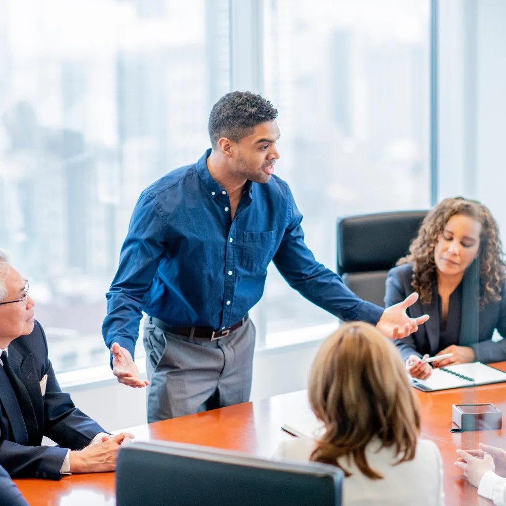 A man in a blue shirt passionately speaks to colleagues around a conference table. Three people listen intently.
