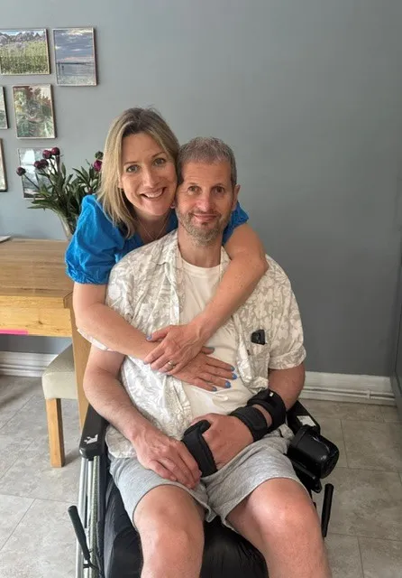 Person sitting in a wheelchair with arm support, embraced from behind by partner at home, reflecting life after stroke and caregiving