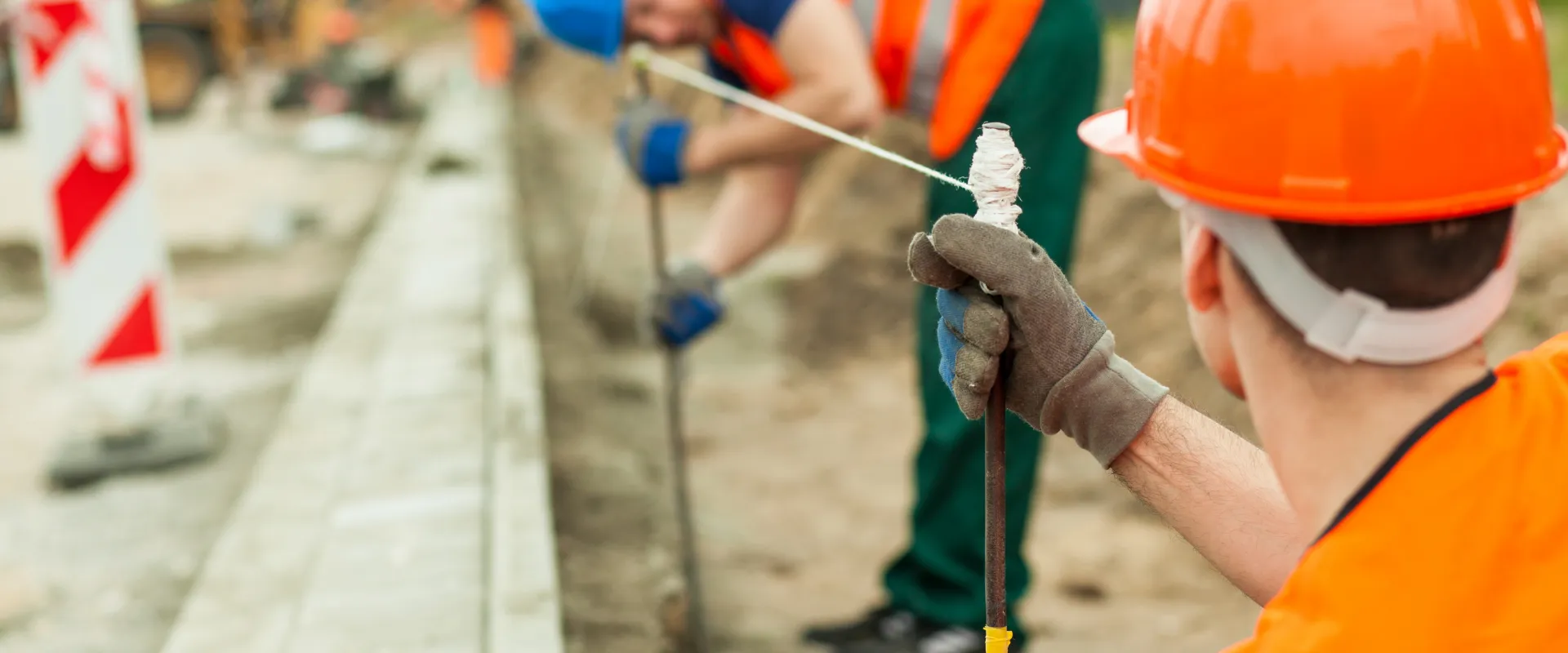 Two construction workers in orange vests and hard hats align a string for measuring on a construction site, focusing intently.