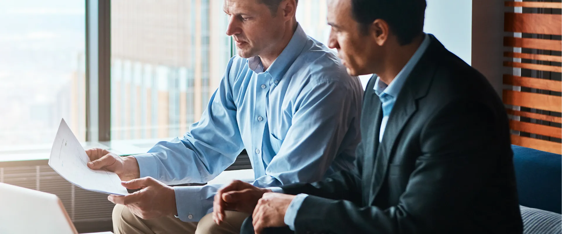 Two men in business attire discuss documents in a modern office, focused and serious. A laptop and papers are on the table.
