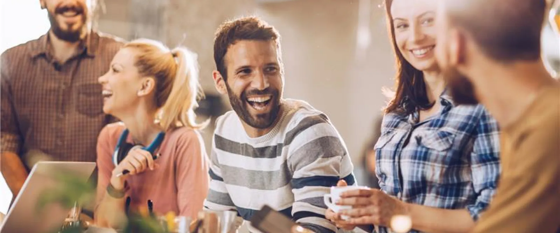A group of five people in a bright office setting share a laugh. They are casually dressed, with laptops and notebooks around.