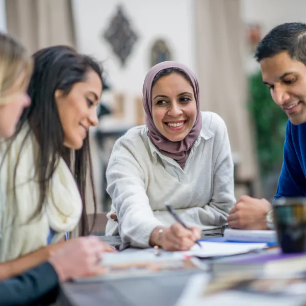 A diverse group of four people sit around a table, engaged and smiling, with papers and books spread out. The mood is collaborative and positive.