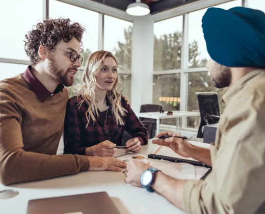 A couple sits at a table, attentively listening to a man with a blue turban. The setting is a bright office with large windows, creating a professional and engaged atmosphere.