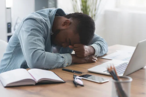 A man in a denim jacket sits at a desk, resting his head on folded arms, appearing tired. An open notebook, phone, and laptop are nearby.