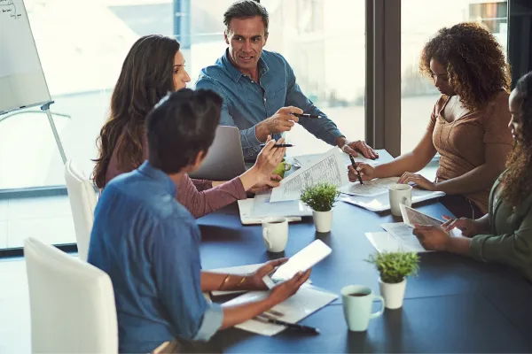 A diverse group of five people in a meeting room sit around a table with notebooks and tablets, actively discussing and pointing at documents.
