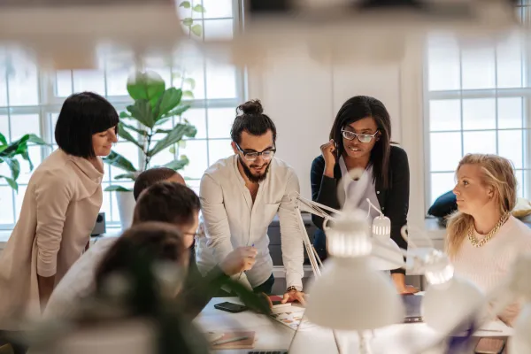 A diverse group of colleagues engaged in a discussion around a table in a well-lit office with plants. They appear focused and collaborative.