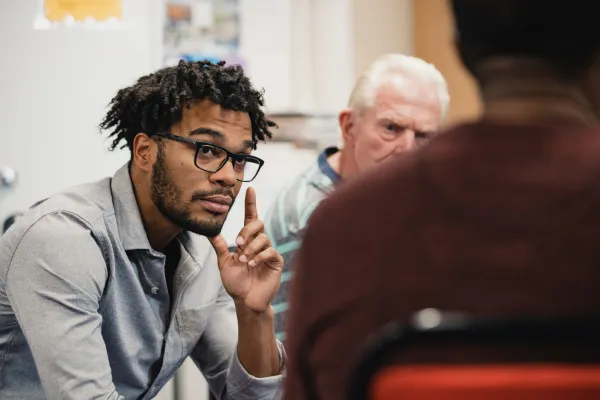A man with glasses attentively listens in a group setting, gesturing thoughtfully. Others in the background suggest a serious yet engaged discussion.