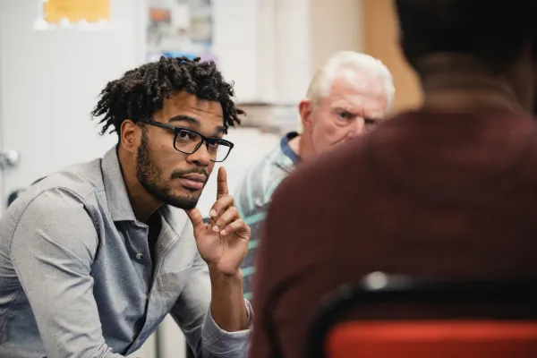 A man with glasses and a thoughtful expression gestures during a group discussion.