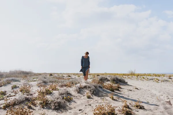 A person in a blue dress walks barefoot on a sandy beach dunes, surrounded by sparse vegetation under a bright, cloudy sky, conveying tranquility.