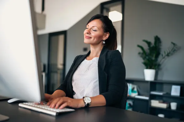 A confident woman in business attire sits at a desk, smiling contentedly with closed eyes. A computer is in front of her.