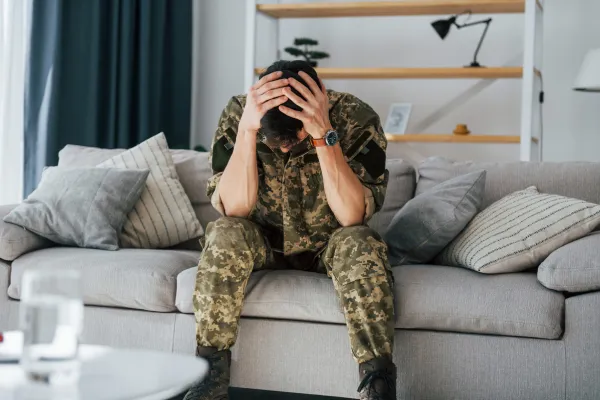 A soldier in camouflage, seated on a couch, with head in hands expressing distress. 