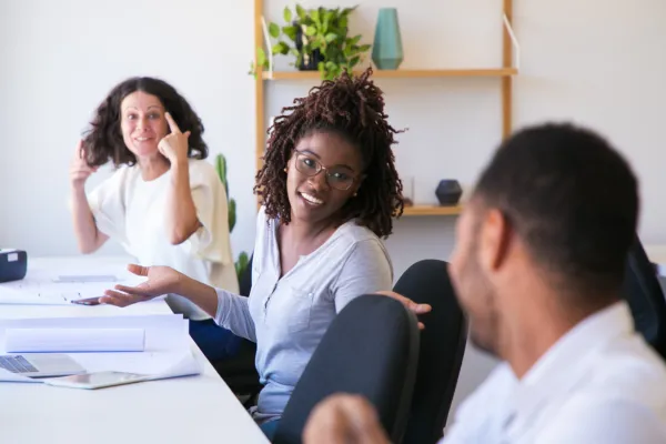 Three colleagues are engaged in a lively discussion at a desk, smiling and gesturing. The background has shelves with plants, creating a casual, friendly office atmosphere.