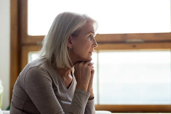 Elderly woman with short white hair wearing a beige sweater, sitting indoors and gazing thoughtfully out a bright window. Calm and reflective mood.