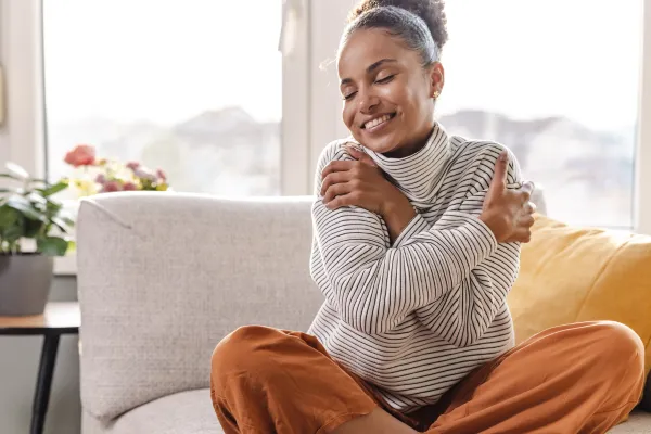 A person sits cross-legged on a light sofa, gently hugging themselves with a content smile. 