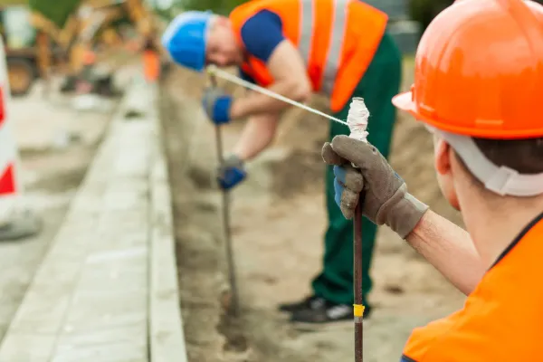 Two construction workers in orange vests and hard hats align a string for measuring on a construction site, focusing intently.