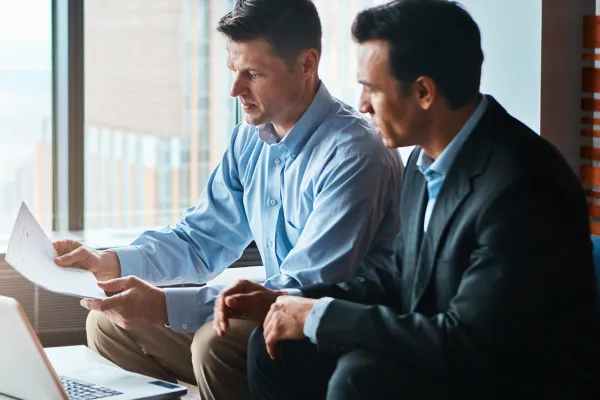 Two men in business attire discuss documents in a modern office, focused and serious. A laptop and papers are on the table.