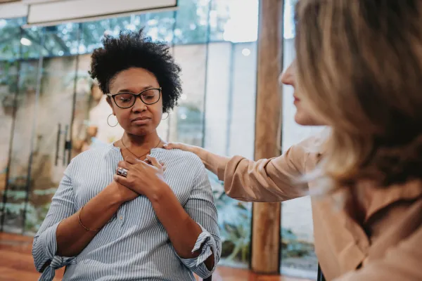 A woman sits with her hand on her chest, looking contemplative. Another person offers comfort with a hand on her shoulder. The scene feels supportive.