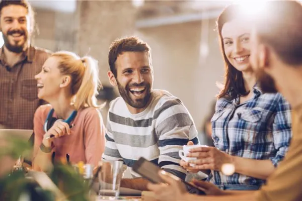 A group of five people in a bright office setting share a laugh. They are casually dressed, with laptops and notebooks around.