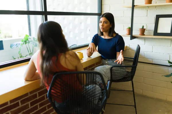 Two women sit at a window-side counter in a café, engaged in conversation.