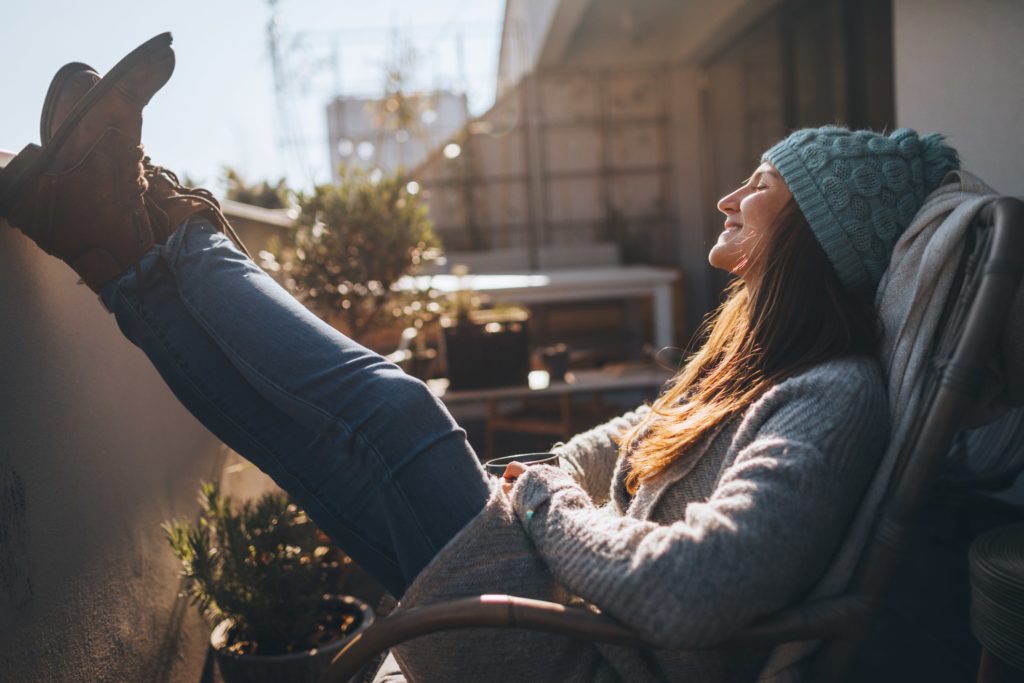 Woman in winter clothes sat outside with feet on balcony smiling