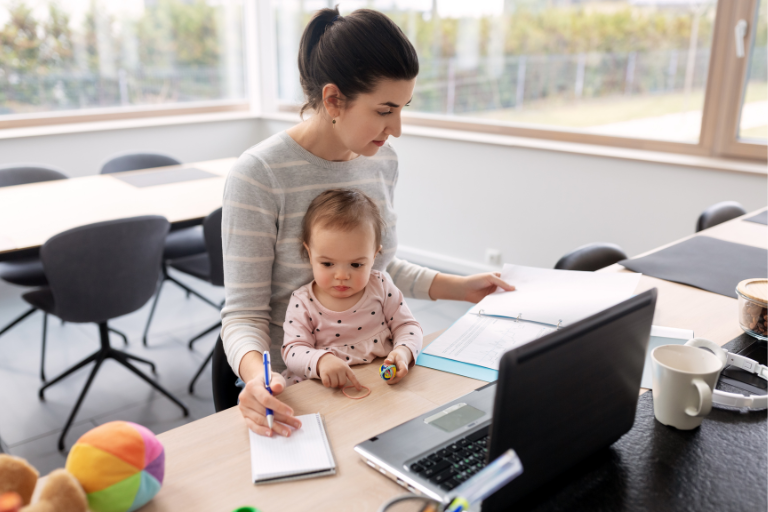 Woman sitting at a desk with a child on her lap while she reads on her laptop.