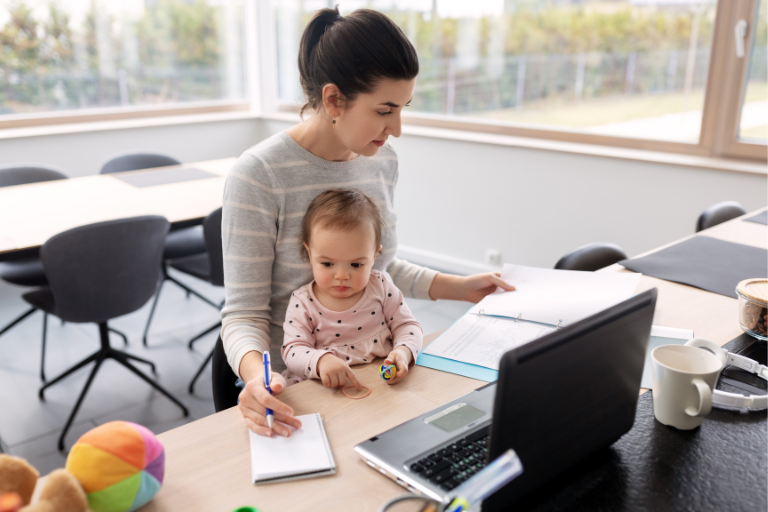 Woman sitting at a desk with a child on her lap while she reads through work papers in front of her laptop