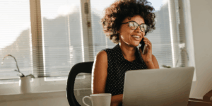 Person sitting at a desk using a laptop and talking on a mobile phone, with a coffee mug nearby and sunlight streaming through window blinds.