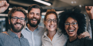 Four people standing indoors, smiling and raising their fists in celebration, wearing glasses and casual clothing.
